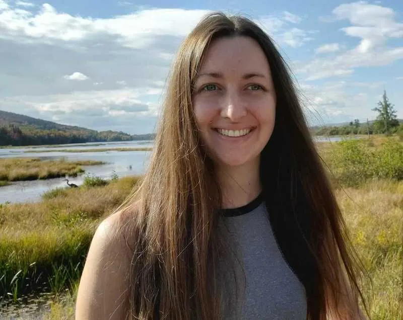 Photo de Vanessa debout devant un paysage naturel avec rivière, herbes hautes et ciel nuageux.