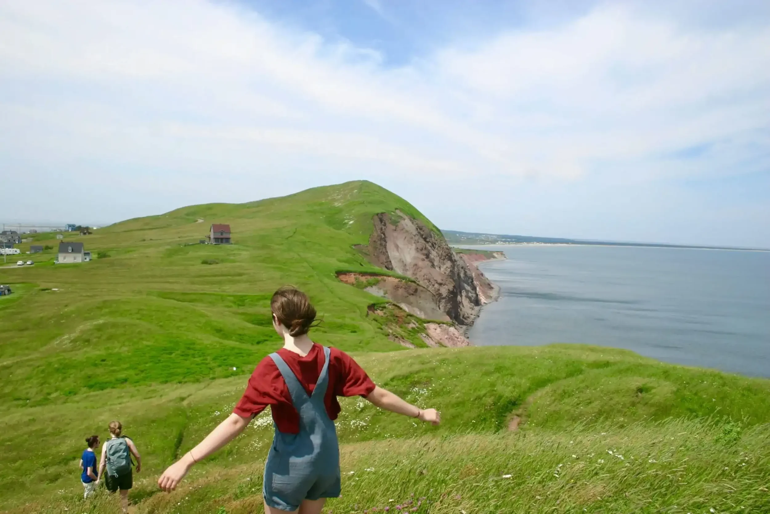 Paysage des Îles de la Madeleine où l'on voit des enfants courir sur les plaines vertes. On voit aussi la mer et des petites maisons colorées.